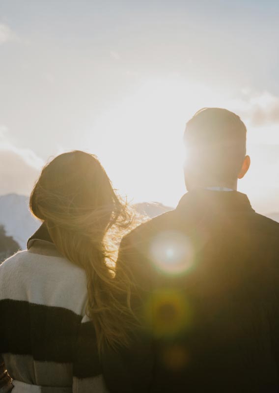Two people look towards the sun setting behind snow-covered mountains.