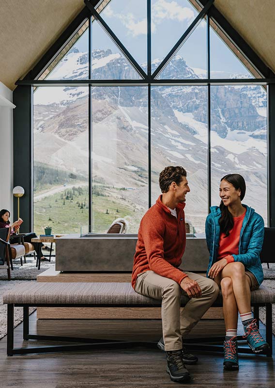 Two people sit in a lounge with a large window overlooking rocky mountain peaks
