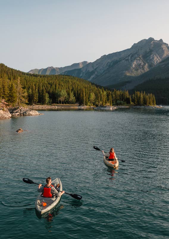 Two kayakers paddle on a blue lake near a forested mountainside.