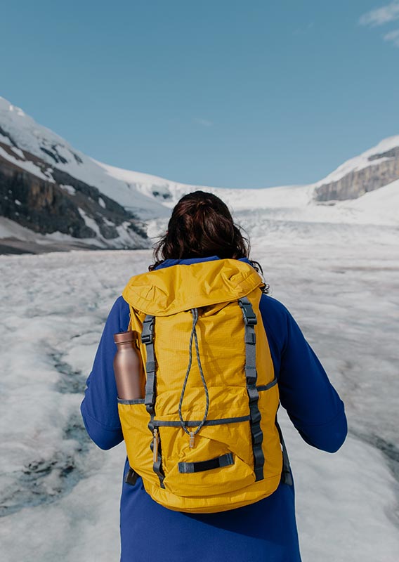 A person in blue jacker and yellow backpack looks out across a glacier between tall mountains.