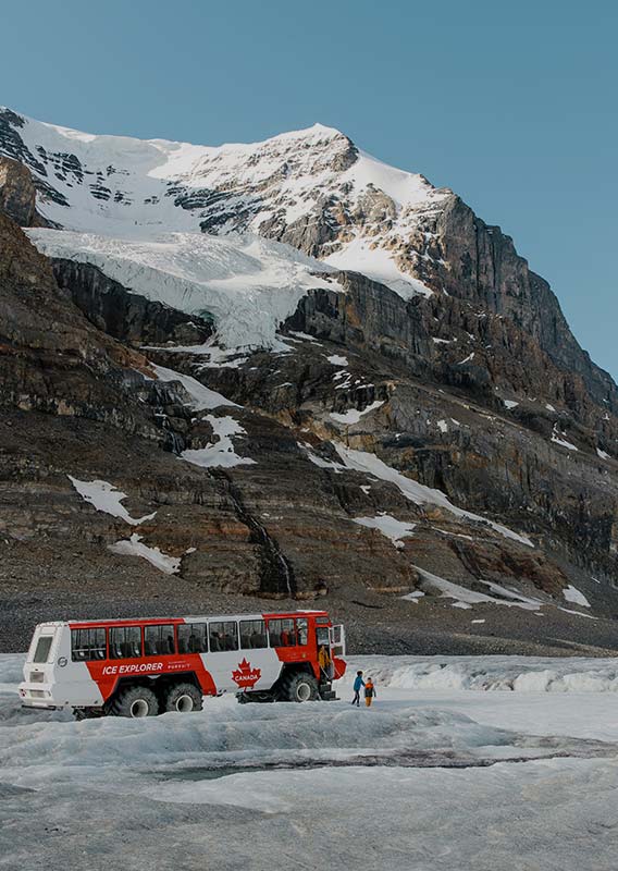A family gets off a large Ice Explorer bus on a glacier, below tall mountains.