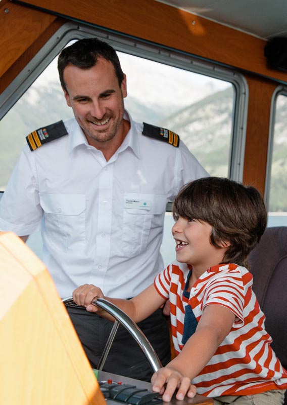 A boy sits at the captains chair of a boat while the captain stands aside