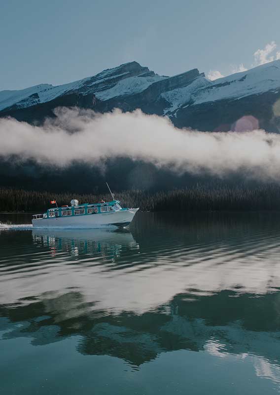 A blue and white boat cruises next to a green and snow-covered mountainside