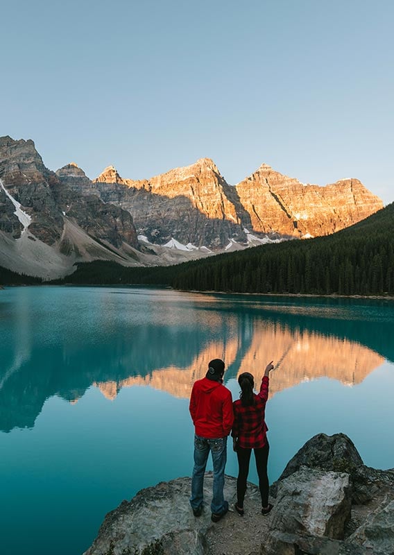 Two people look out over a blue lake towards towering mountain peaks.