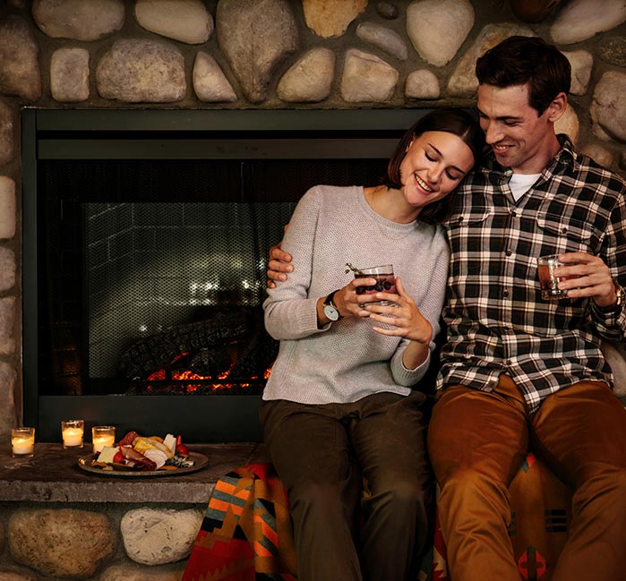Two people relax by a fireplace, sitting with drinks and a charcuterie board.