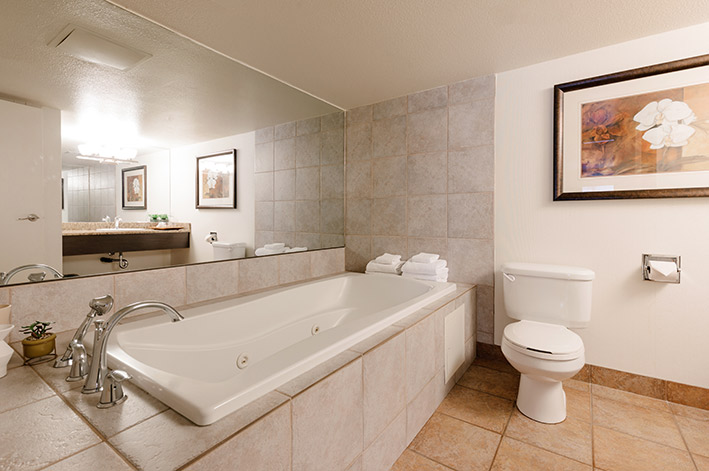 A tiled jetted tub with towels next to a toilet and large mirror