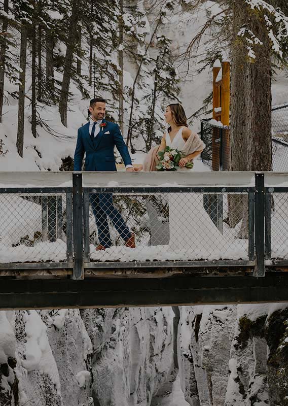 A newlywed couple walks across a bridge over a snowy and icy canyon.