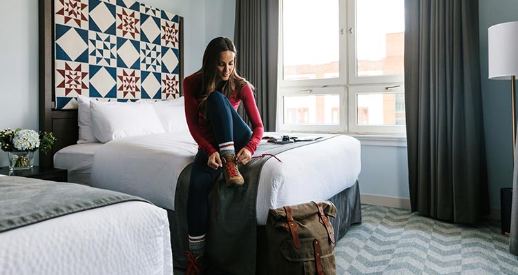 A woman sits on a hotel bed, putting on hiking boots.