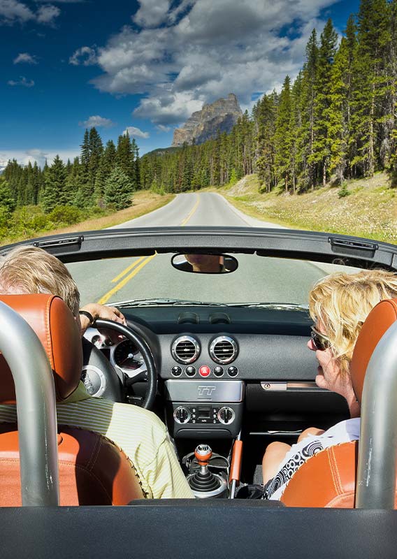 Two people driving a convertible car down an open road between forests.