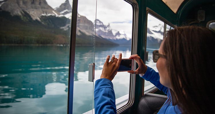 A person holds a camera up to a boat window to take a photo of a mountain landscape.