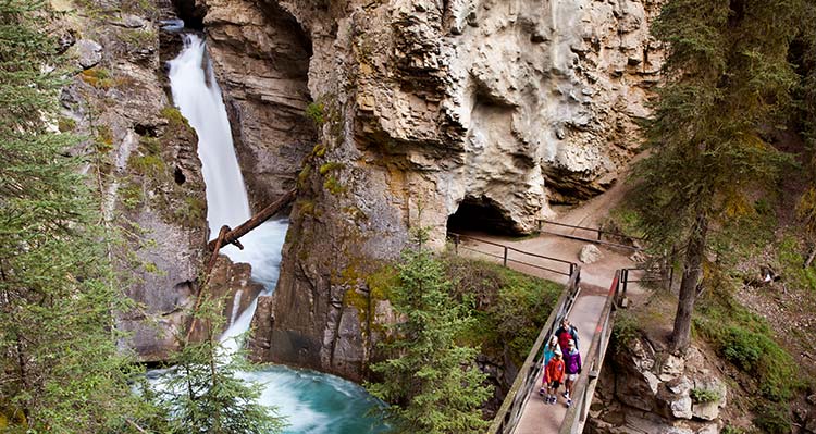 A family walks across a little bridge over a canyon near a rushing waterfall.