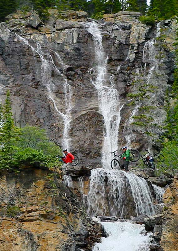 Three mountian bikers walk on a path below a rushing waterfall.