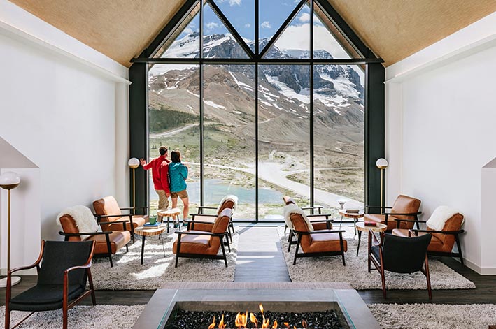 A lounge with a fireplace next to a large pointed window to a view of mountains and a glacier