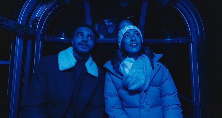 Two people ride a gondola cabin, lit up with blue light.