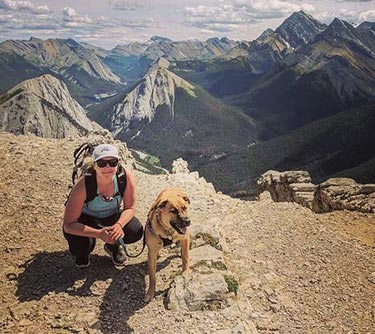 A woman crouches with a dog at the top of a mountain.