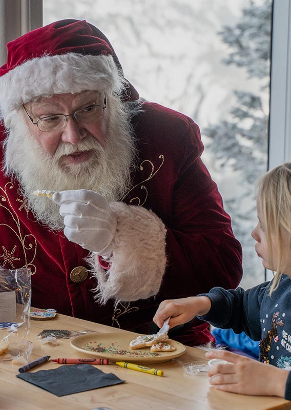 Santa sits at a table with children decorating cookies