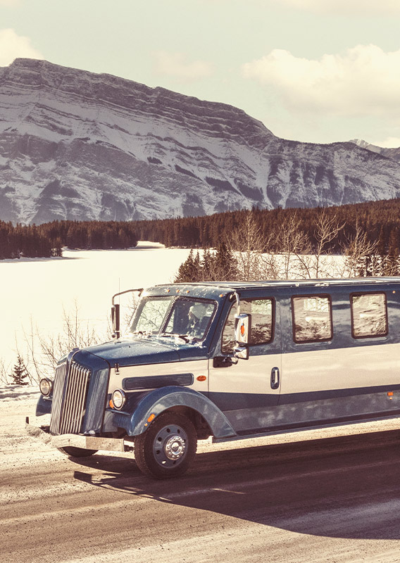 A historic style bus drives from a bridge away from a small town, with mountains in the background.