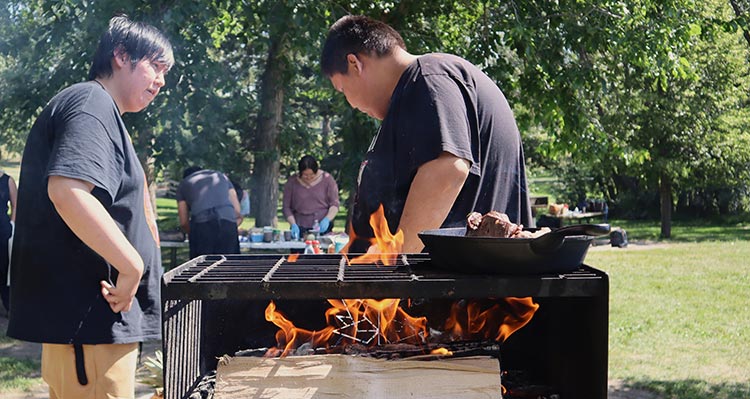 Two people cook together over a wood-fired grill  in a park.