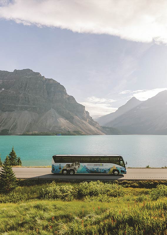 A Brewster Express bus drives along a road next to water, trees and mountains