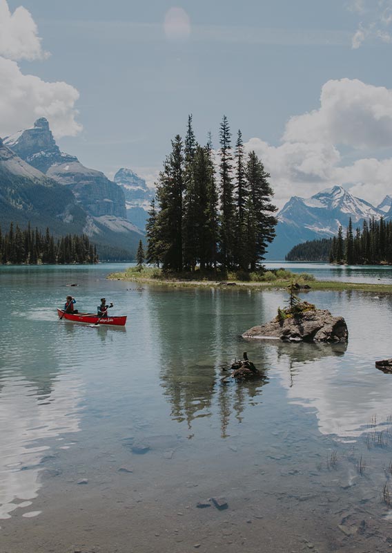 Two canoests paddle on lake near a small tree-covered peninsula.