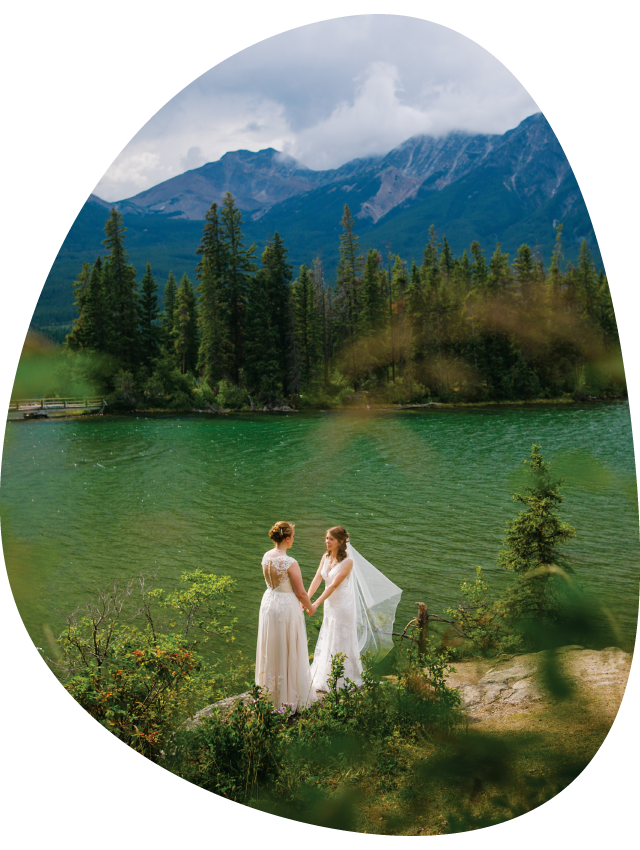 Two brides hold hands at a rocky viewpoint above a small mountain lake.